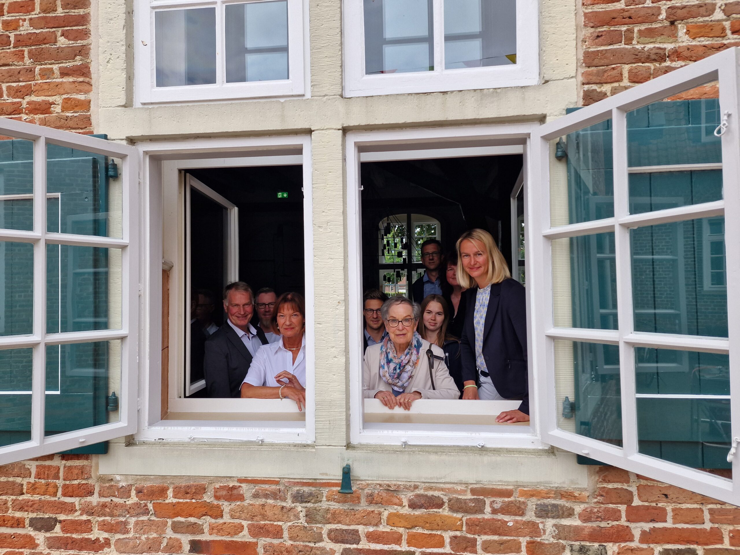 Gruppe von Menschen schaut lächelnd aus zwei offenen Fenstern eines roten Backsteingebäudes, vorn ältere Frau mit Halstuch.