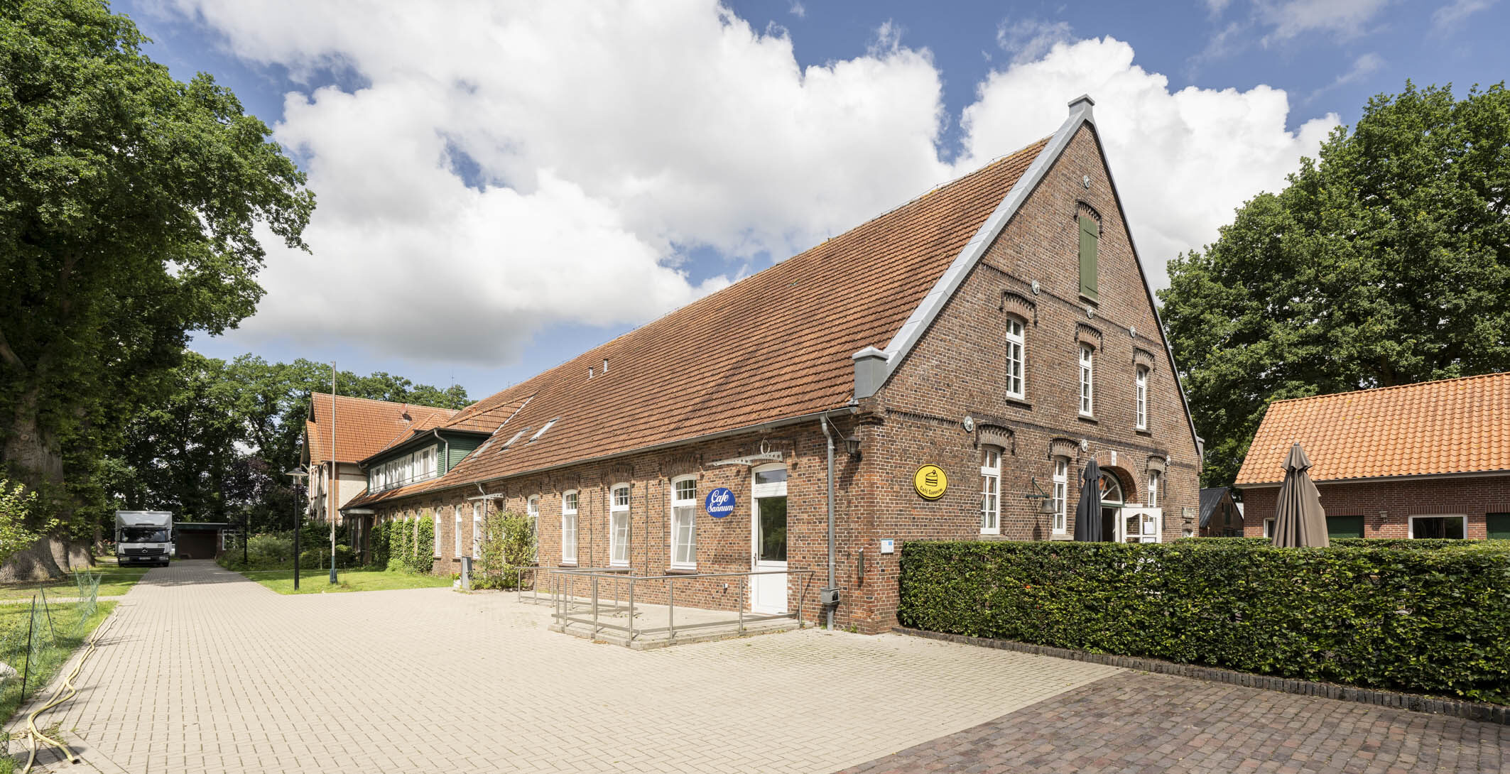 Hof mit roten Backsteinhäusern, hölzerner Kinderschaukel, Bänken, Kopfsteinpflaster und Bäumen hinter weißem Tor.