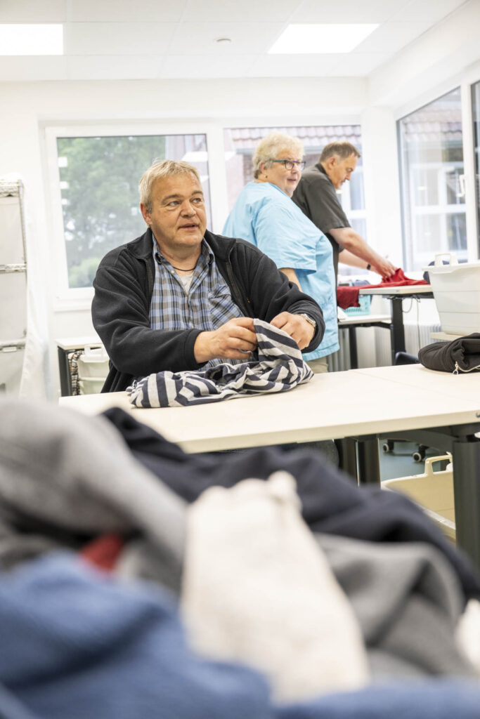 Älterer Mann sitzt an einem Tisch und faltet gestreiftes Shirt, im Hintergrund zwei Personen beim Wäschefalten.