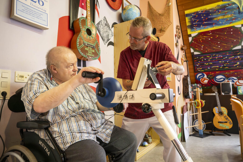 Mann im Rollstuhl übt an einem Armkurbelgerät, ein Therapeut in roter Kleidung hilft ihm, im Hintergrund bunte Gitarren und Wandkunst.