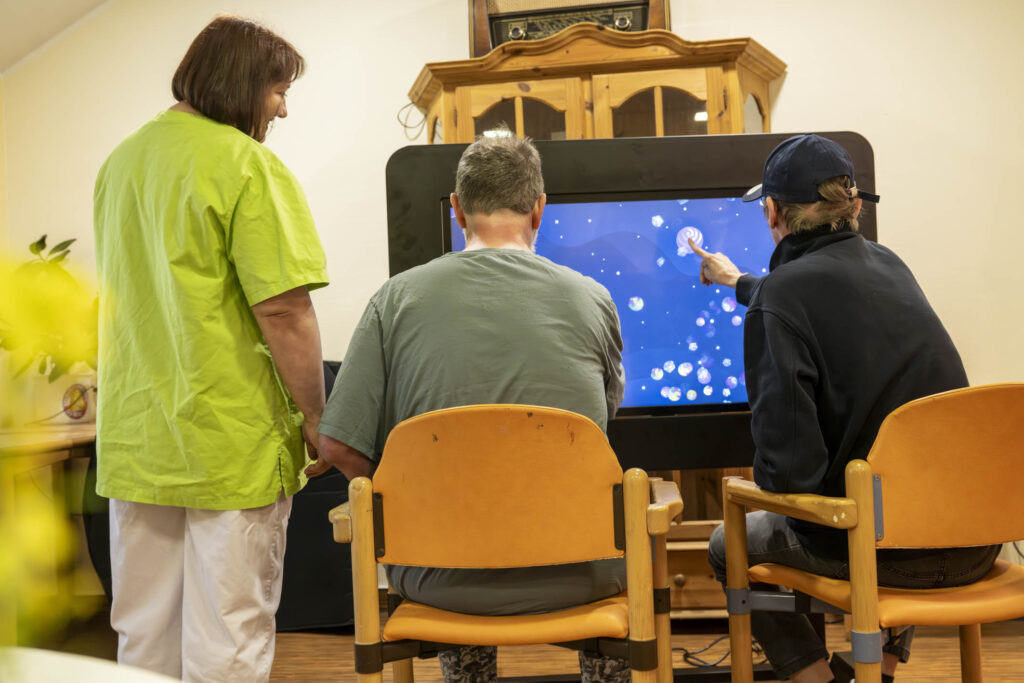 Pflegekraft in grüner Dienstkleidung steht neben zwei Männern, die auf einem großen Touchscreen mit blauen Blasen interagieren.