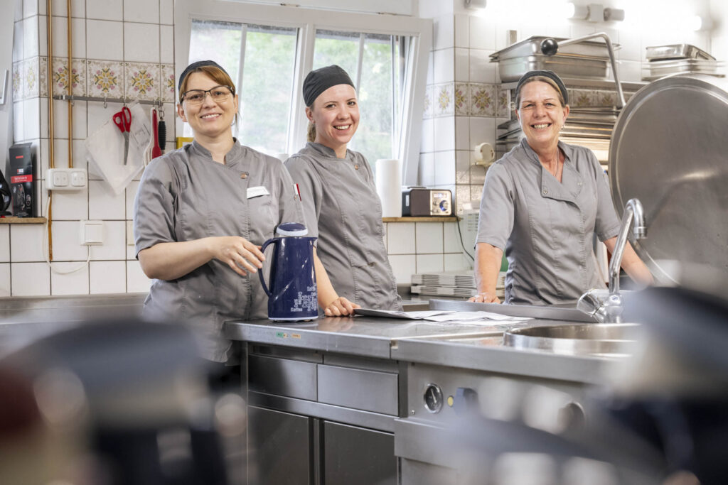 Drei lächelnde Köchinnen in grauen Kitteln stehen an einem Edelstahltisch in einer Großküche; vor ihnen steht eine blaue Kanne.