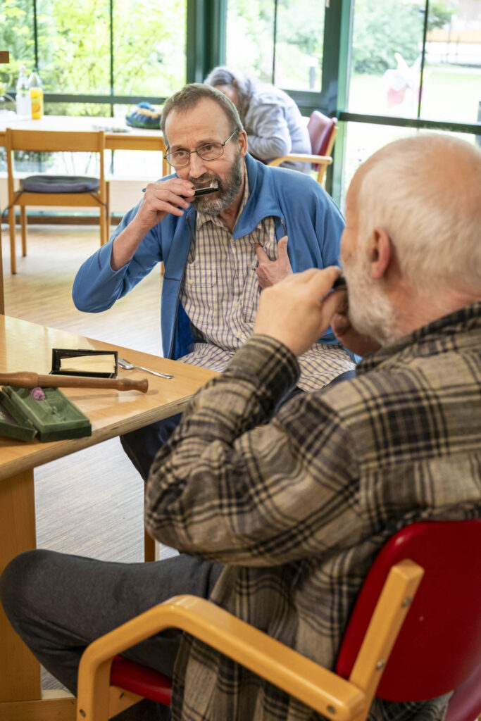 Zwei ältere Männer in einem Aufenthaltsraum spielen Mundharmonikas; einer mit Brille und blauem Jackett schaut nach vorn.
