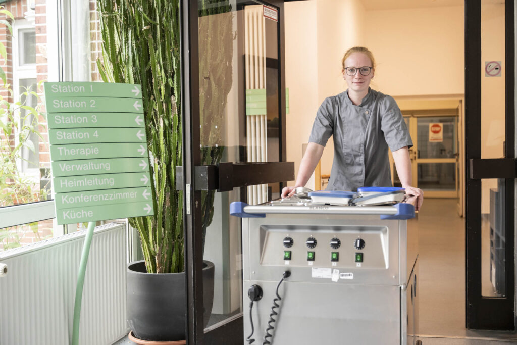 Mitarbeiterin in grauer Dienstkleidung schiebt einen Edelstahlwagen durch den Eingang eines Pflegeheims, links ein grünes Wegweiser-Schild.