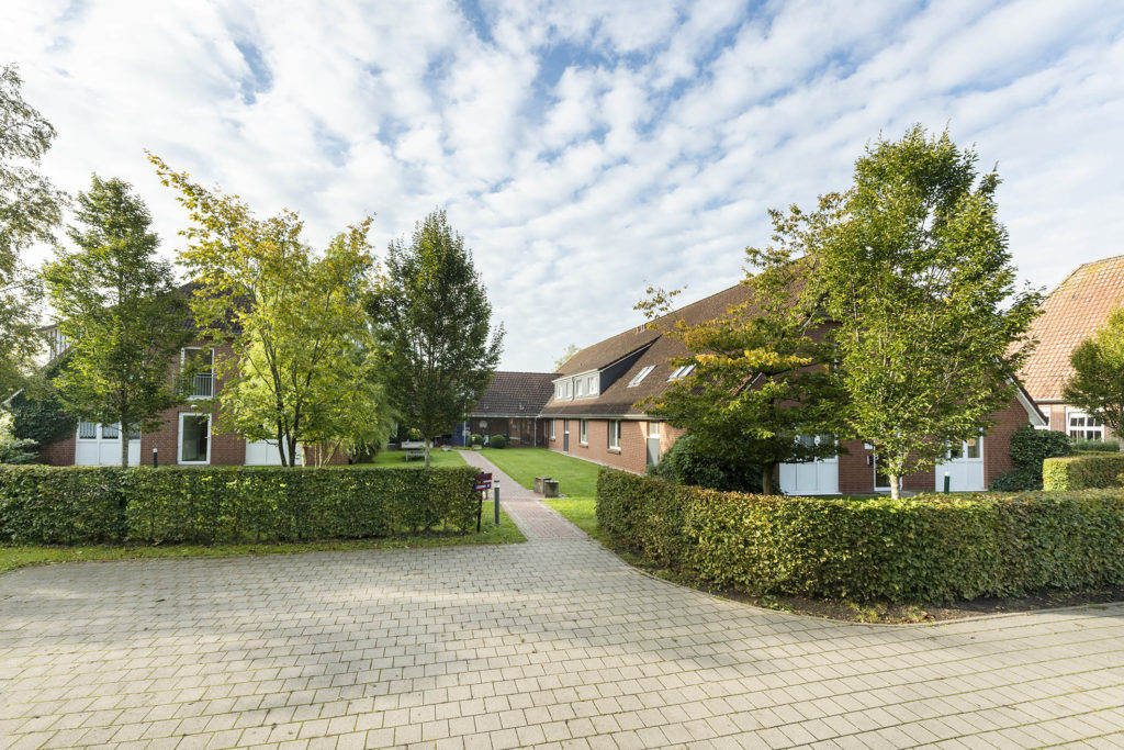 Innenhof mit roten Backsteinhäusern und schrägen Ziegeldächern, Bäumen, gepflastertem Weg zwischen Hecken und leicht bewölktem Himmel.