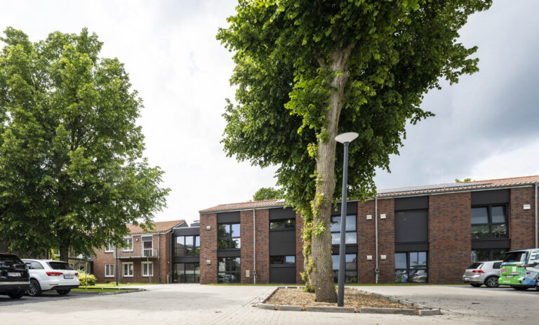 Eingangsbereich eines zweistöckigen Gebäudes mit roten Ziegeldächern, Glasvordach, gepflastertem Weg und Bäumen.