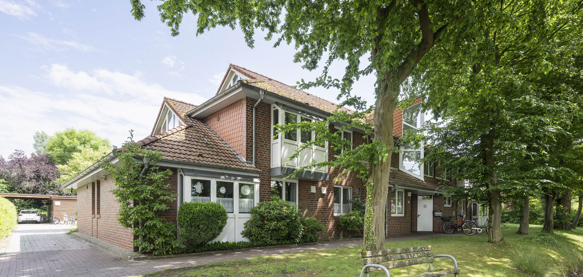 Rotes Backstein-Reihenhaus mit Ziegeldach in grüner Anlage, Bäume und Parkbank im Vordergrund und Fahrräder am Eingang.