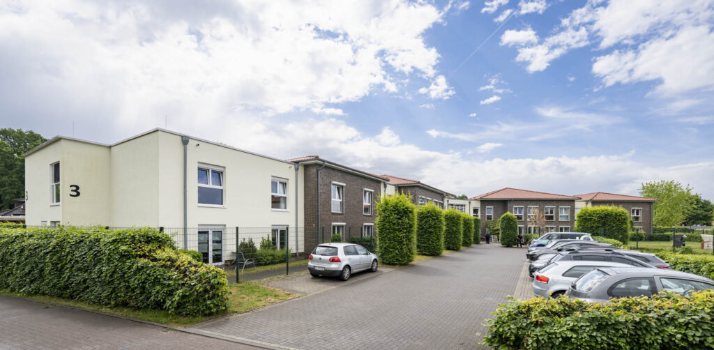 Zweistöckiges Wohngebäude mit roten Ziegeldächern, Rasenfläche im Vordergrund, parkenden Autos und bewölktem Himmel.
