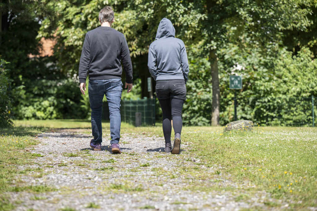 Zwei Personen in Freizeitkleidung, eine mit Kapuzenpullover, gehen auf einem Kiesweg durch einen grünen Park.