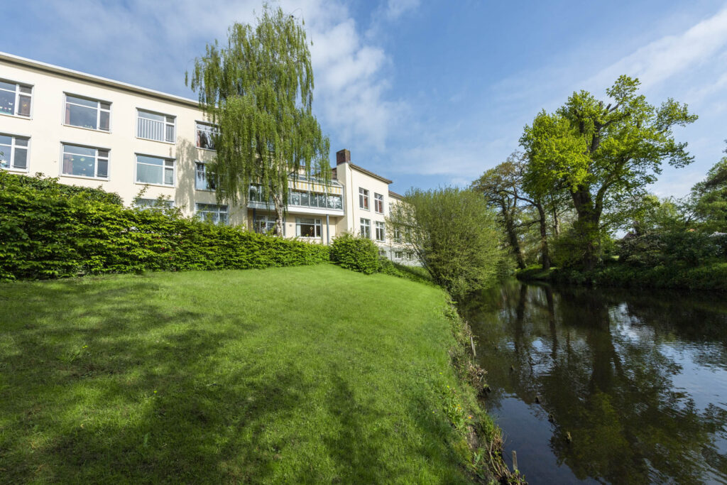 Pferde-Skulptur auf Rollen auf grünem Rasen vor einem Gebäude, weiße Rhododendronblüten links und rotlaubiger Baum rechts.