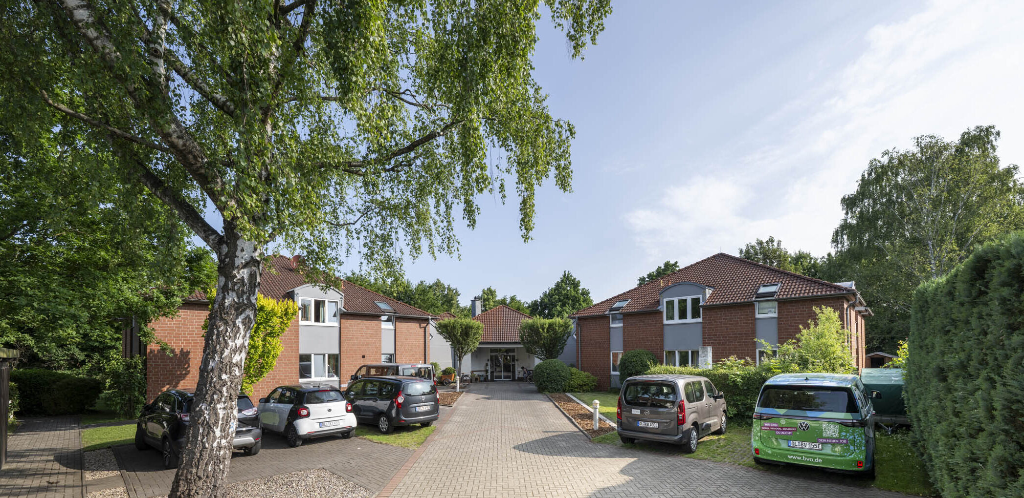 Hof mit gepflasterter Einfahrt, roten Backsteinhäusern, geparkten Autos und einer großen Birke vorne.