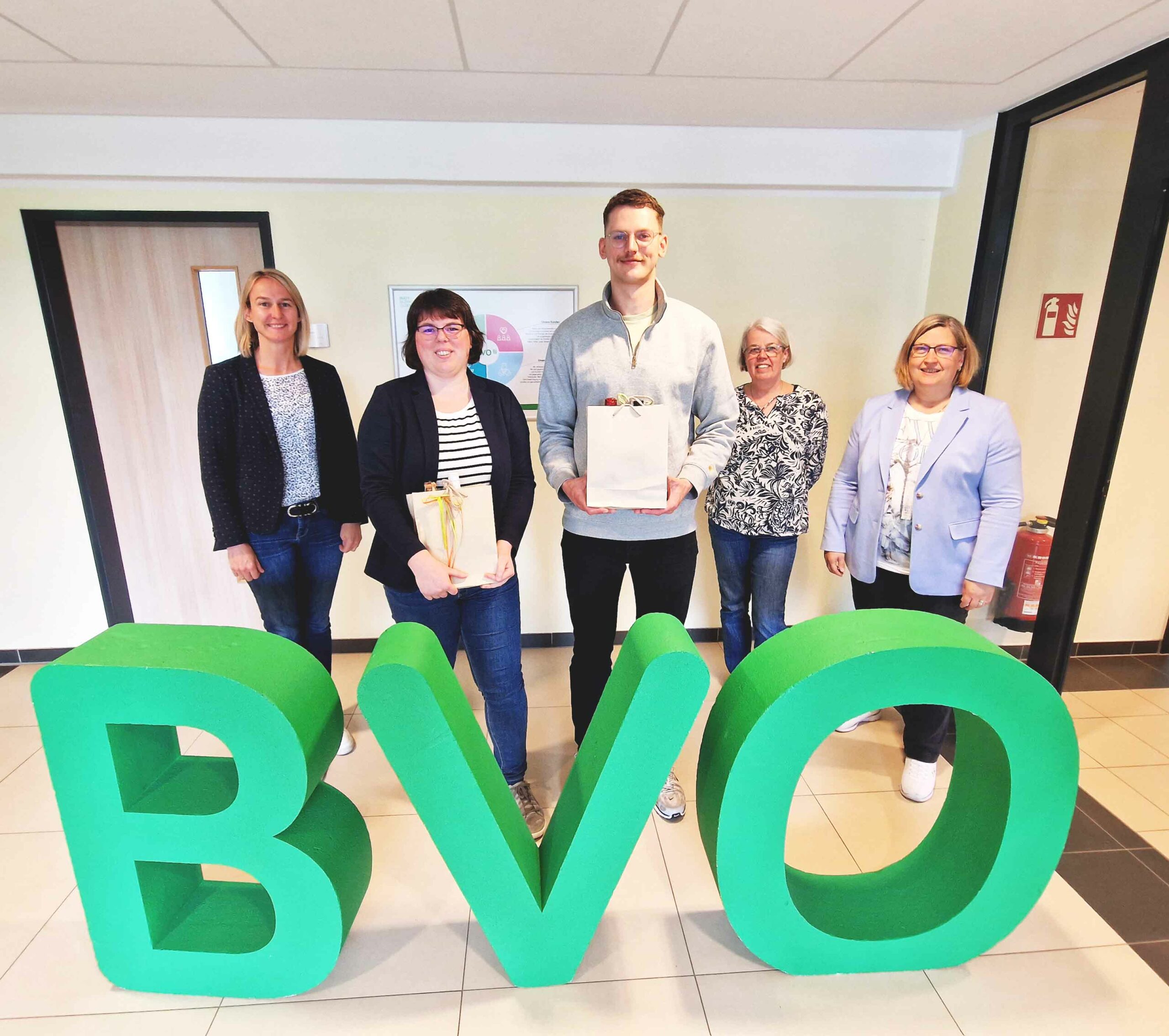 Fünf Personen stehen in einem Büroflur hinter großen grünen Buchstaben BVO, zwei von ihnen halten Geschenkbeutel.