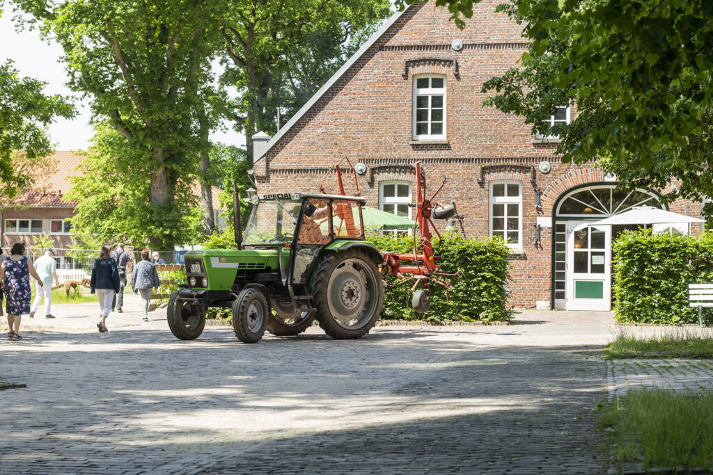 Grüner Traktor mit rotem Anbaugerät auf Kopfsteinpflaster vor einem Backsteinhaus, Spaziergänger im Hintergrund.