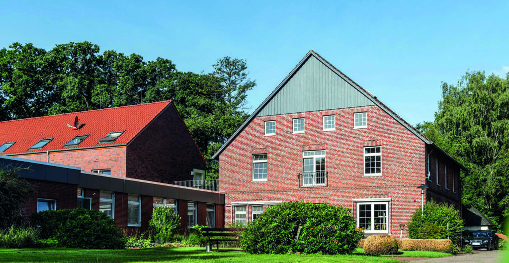 Reihen von Backsteinhäusern mit roten Ziegeldächern, Rasenflächen, Sträuchern und gepflastertem Vorplatz unter klarem blauem Himmel.