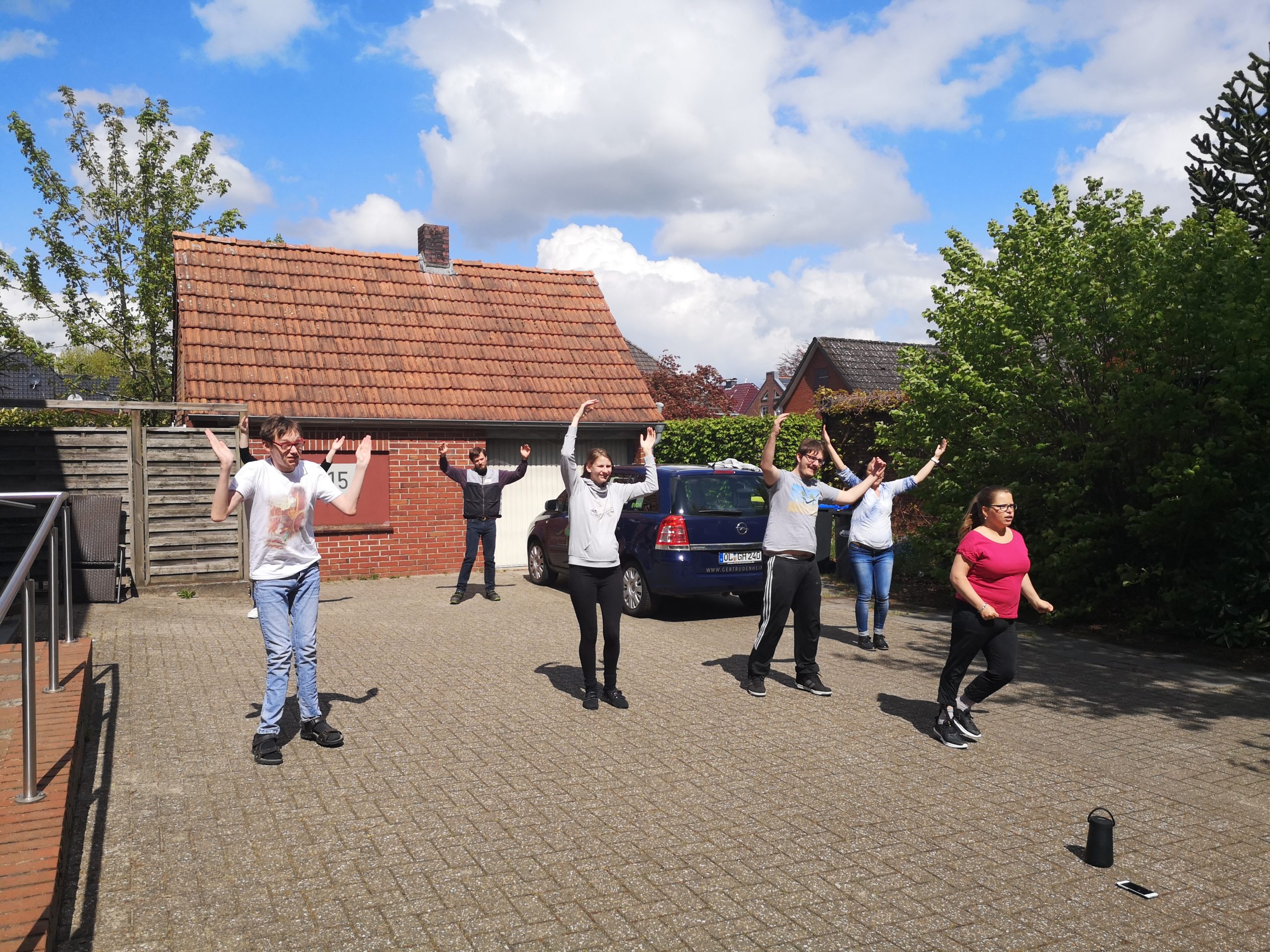 Sechs Personen auf gepflastertem Hof vor einem Backsteinhaus heben die Arme und machen gemeinsam Übungen.