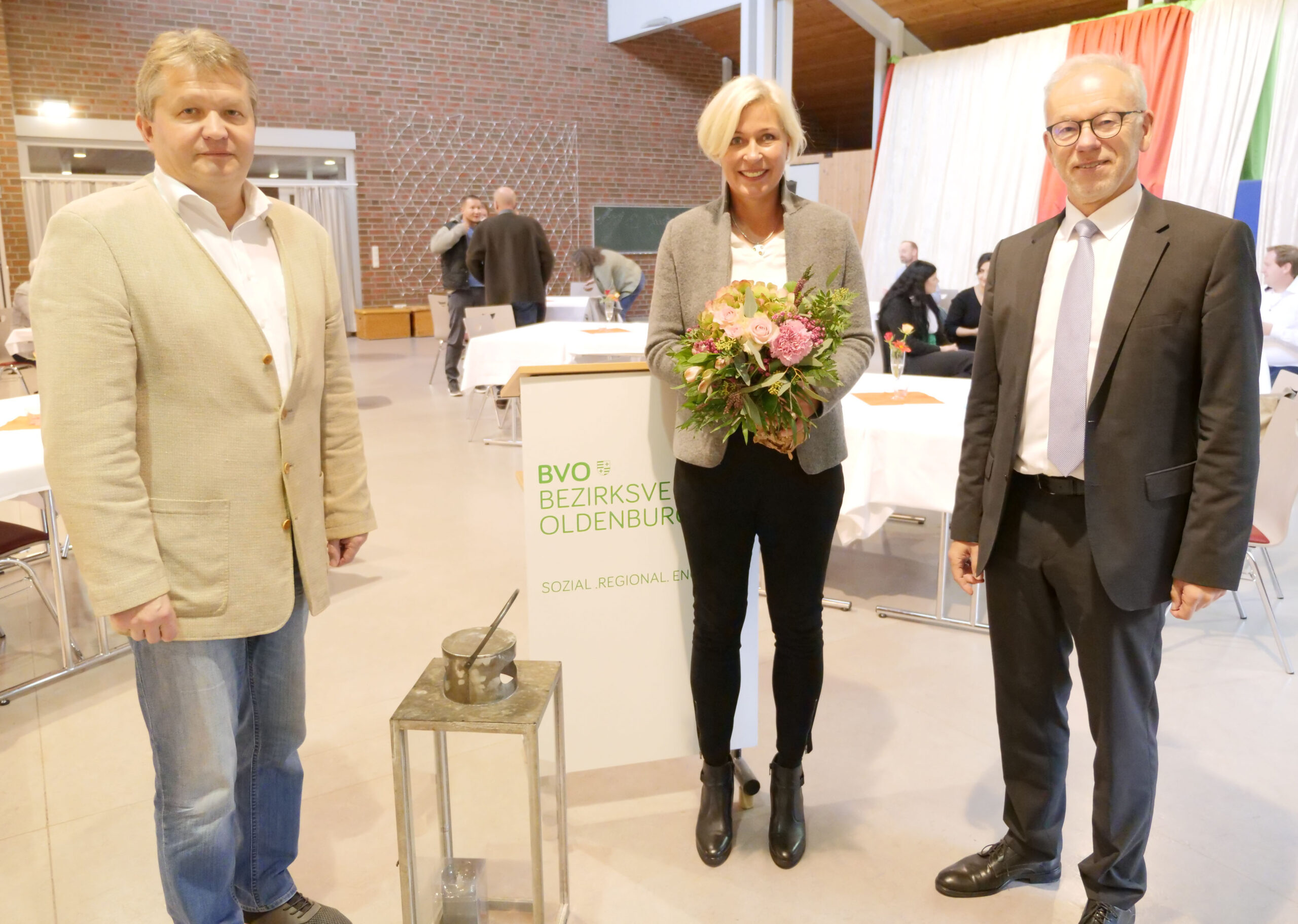 Drei Personen in einer Halle; die Frau in der Mitte hält einen Blumenstrauß, zwei Männer stehen links und rechts vor einem BVO-Infostand.