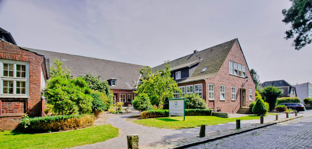 Rotes Backsteingebäude mit steilem Ziegeldach, Vorgarten, gekiestem Weg und Infotafel am Eingang