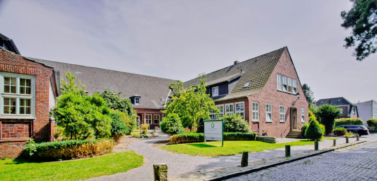 Rotes Backsteingebäude mit steilem Ziegeldach, Vorgarten, gekiestem Weg und Infotafel am Eingang